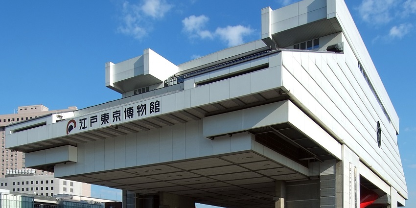 An image of a modern angular building, white walls, a parking area and a clear sky.
