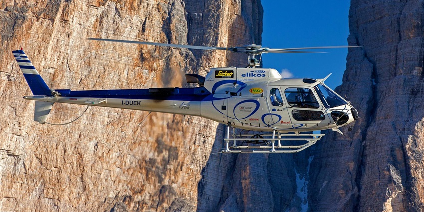A white helicopter flying over rugged brown mountain terrain under a clear sky.