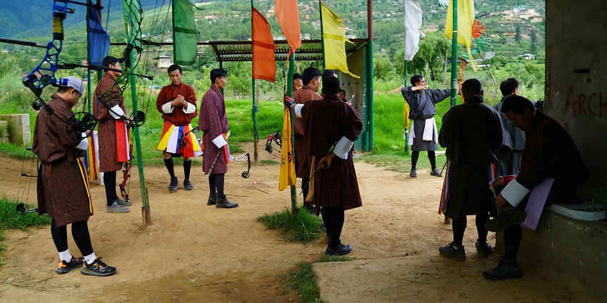 An image of two archers in action, one aiming a compound bow, colourful flags behind them