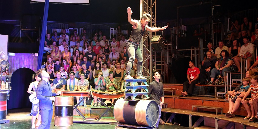 An image of a skilled performer balancing on stacked boards, metal cylinders, a drum, and a tire.