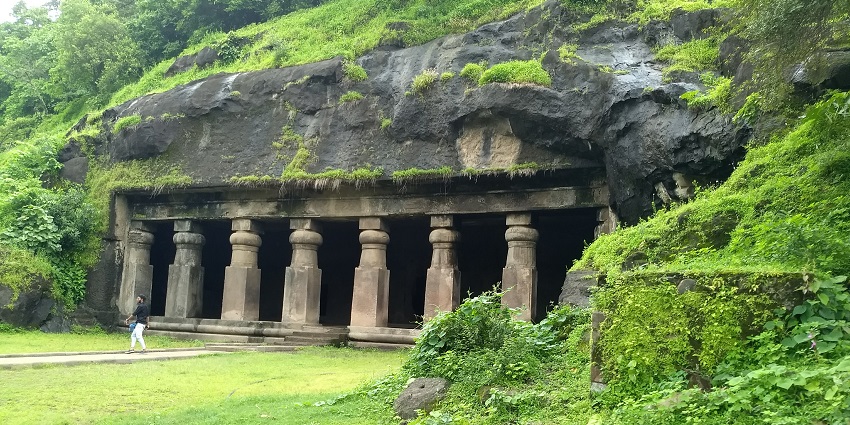 An image of the rock-cut Elephanta caves with large, intricately carved stone pillars set into a green hillside
