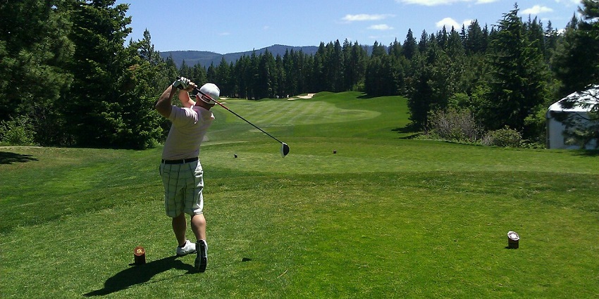 Golfer taking a swing on a sunny day at a scenic golf course surrounded by tall trees.