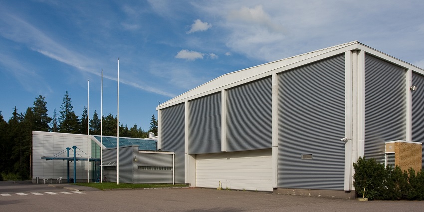 An image of the Finnish Aviation Museum building with corrugated metal exterior, flagpoles, greenery, and clear sky.