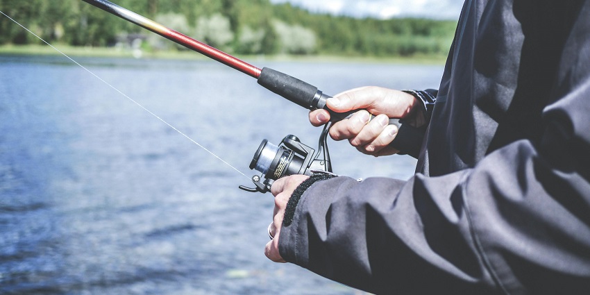 Person holding a fishing rod, reeling the rod in a lake and green mountains in the back