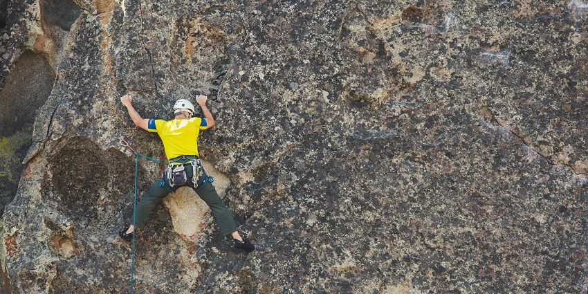 Rock climber in a yellow shirt scaling a rugged cliff, secured with safety gear and rope.