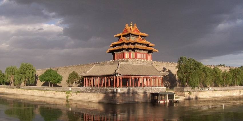 A corner tower of the Forbidden City is standing beside a moat under a cloudy sky.