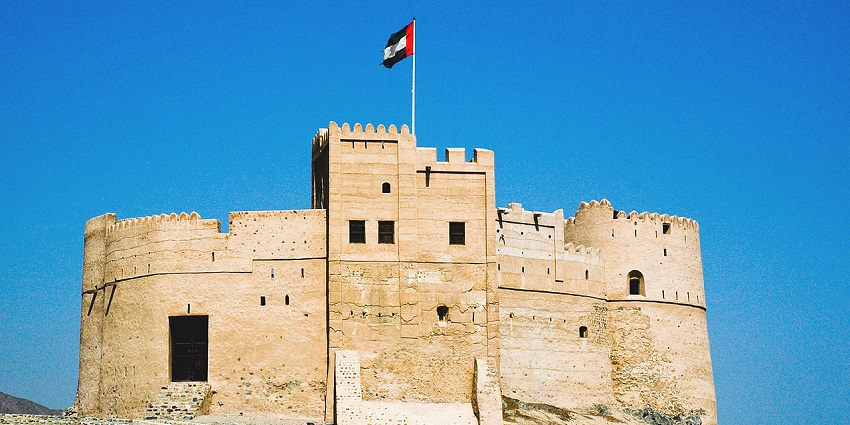 Historic beige fortress with an Emirati flag flying on top, set against a clear blue sky.