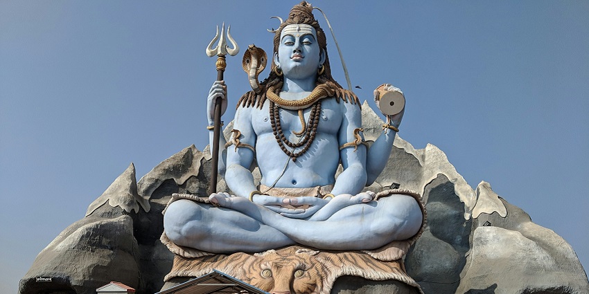 A light-blue statue of Shiva in a seated pose, with a smaller fenced structure in front.