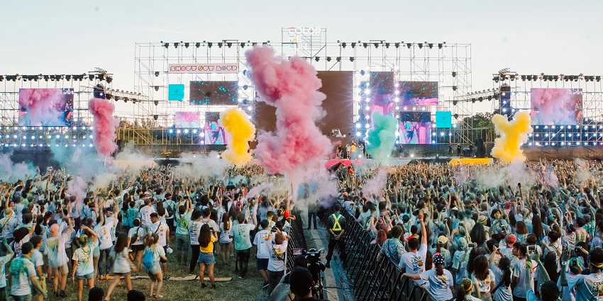 A large crowd at an outdoor festival with colourful smoke and a stage in the background.