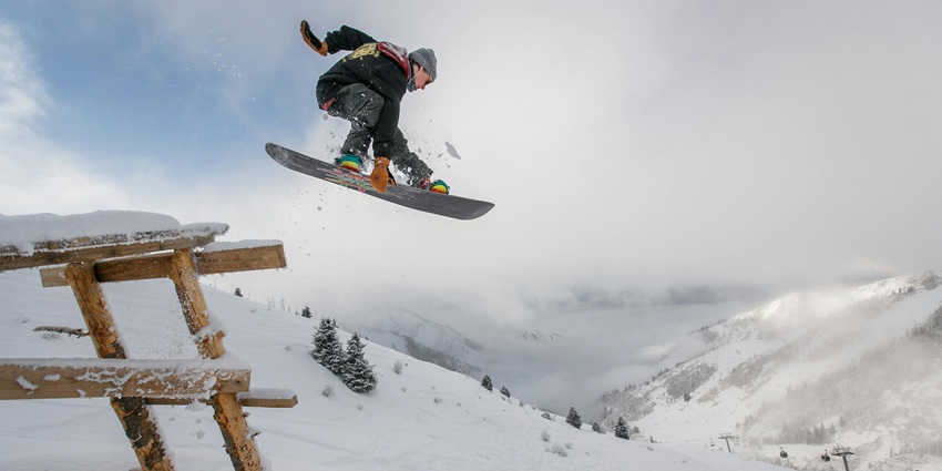 Snowboarder in black gear mid-air performing a jump against a snowy mountain backdrop