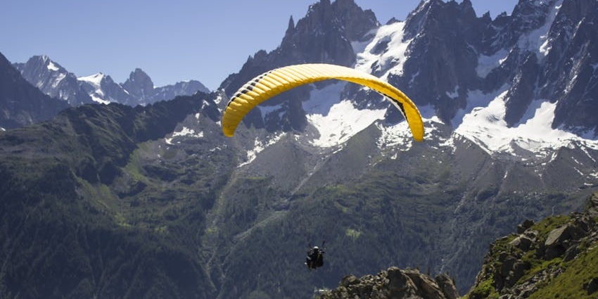 An image of a person paragliding with a yellow paraglider over a green mountainous landscape.