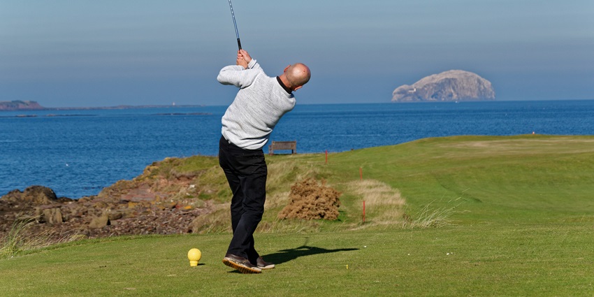A golfer is swinging his club while watching the ball soar over the scenic coastline.