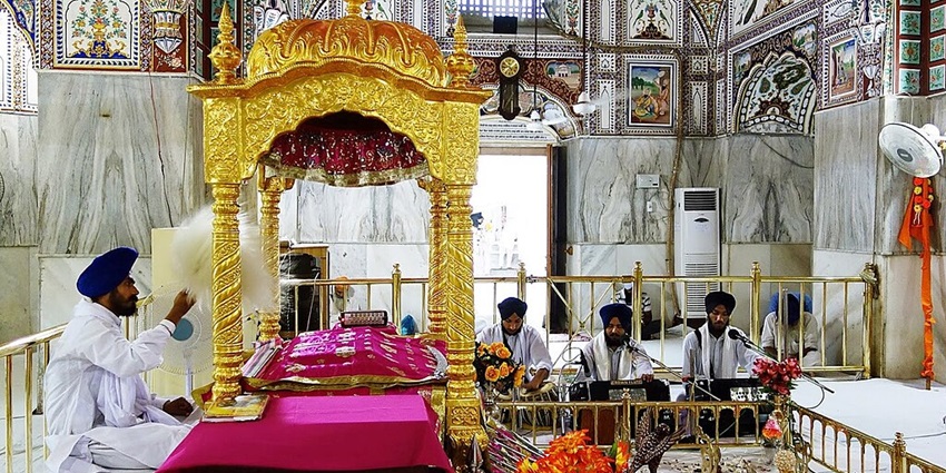 Inside a gurudwara with three men sitting with harmonium and another on a platform.