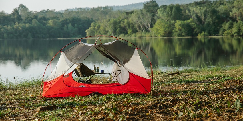 Tent on a shore with a small table and chair inside the tent, and lake surrounded by trees