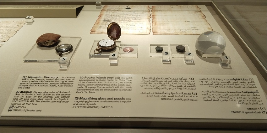 Display featuring Qawasim currency, a replica pocket watch, and a magnifying glass.