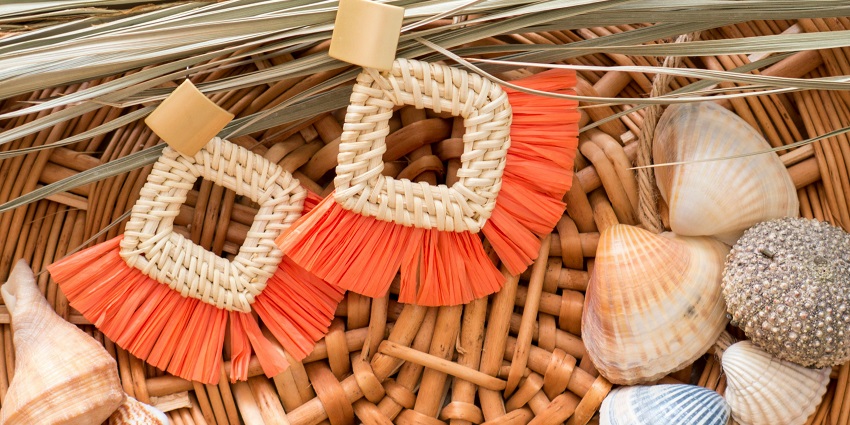 Woven earrings with orange fringe, placed on a wicker surface surrounded by seashells