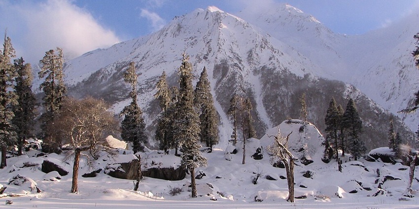 An image of snow-covered mountains with tall trees scattered among large rocks and boulders in the Har Ki Dun Trek, Uttarakhand.