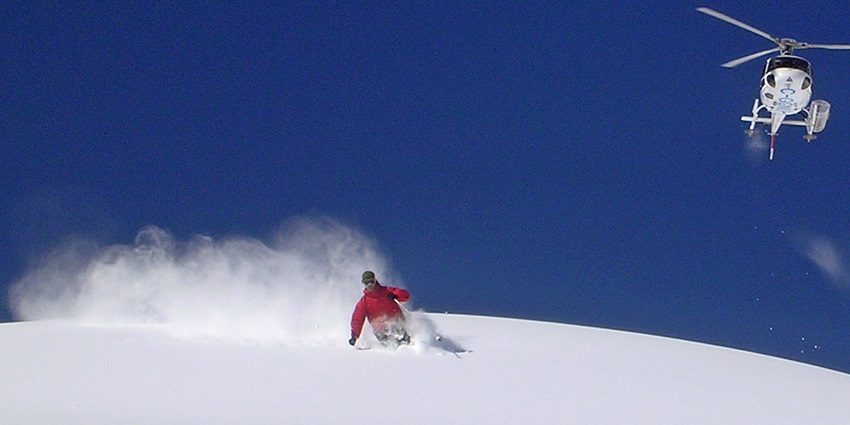 A skier is gliding down a snowy slope while a helicopter is hovering in the clear sky.
