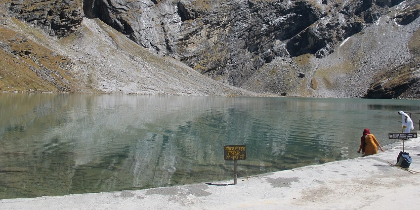 Hemkund Lake with a clear water body, sign boards, two women standing and snowy mountains