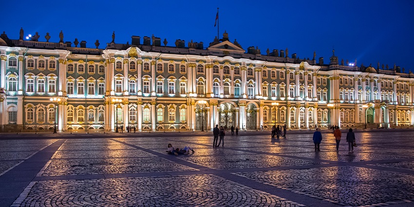 Night view of the Hermitage Museum in St. Petersburg with warm lights illuminating the Winter Palace facade and people walking in the foreground.