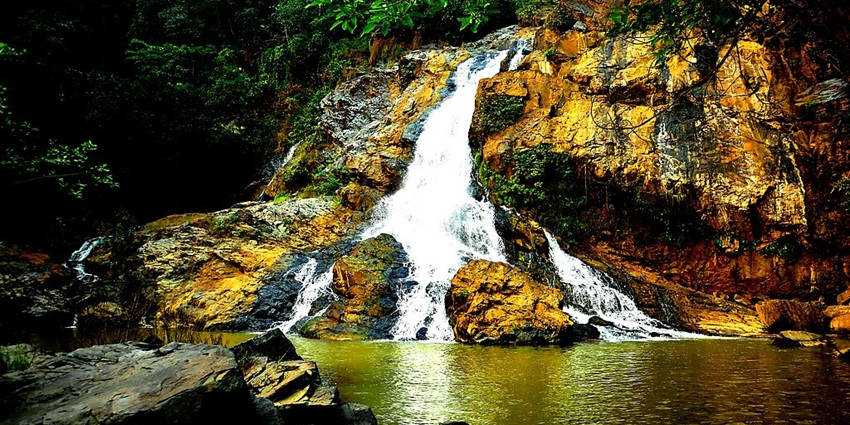 Hirni Falls flows down rocky terrain into a lake, surrounded by dense green forests.