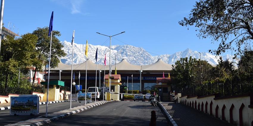 The entrance of Kangra Airport Gaggal with flags, lush trees, and mountains in the background