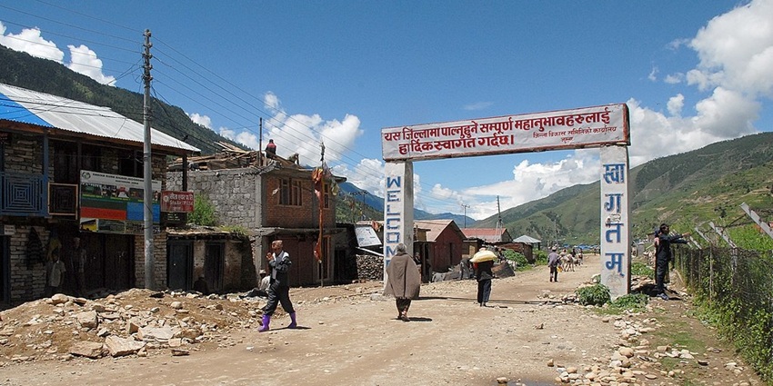 A road in Jumla with a signboard, people walking, small buildings and green mountains.