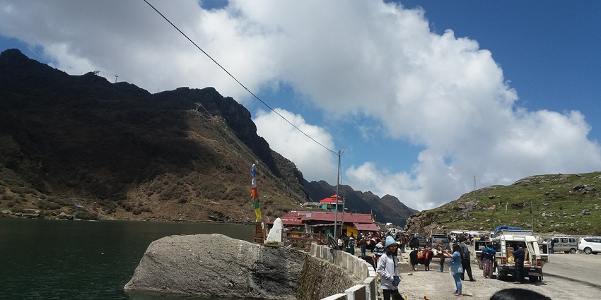 People standing and taking pictures near Changu lake with vehicles on the road and mountains