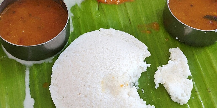 An image of Idli Sambhar, with two idlis, sambar, coconut chutney, tomato chutney, and mint chutney on a banana leaf