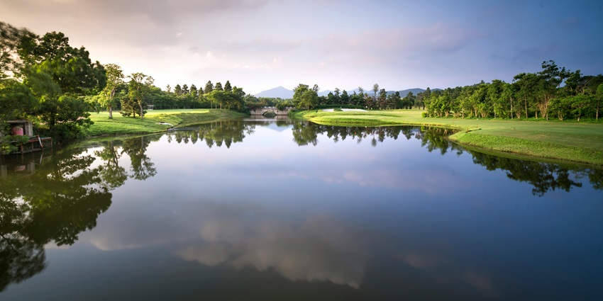 A landscape featuring a calm reflective lake, lush green golf course, and a cloudy sky.