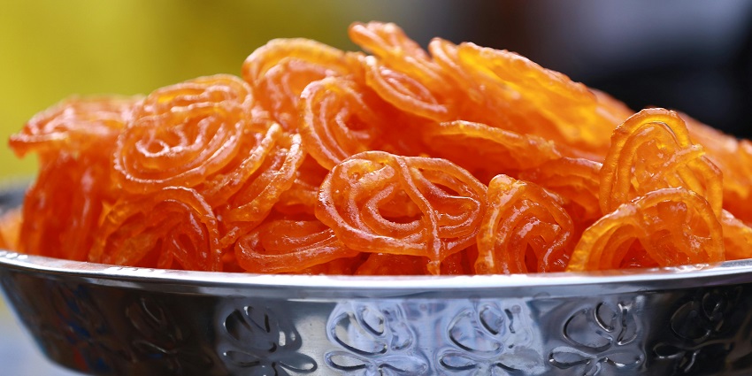 An image of intricate, orange-coloured jalebi spirals glistening with sugar syrup, stacked and ready to serve