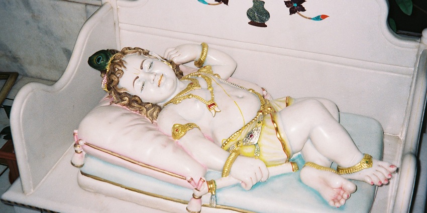 An image of the detailed statue of a reclining baby Krishna, adorned with gold jewellery on a decorated bed, during the Janmashtami festival.