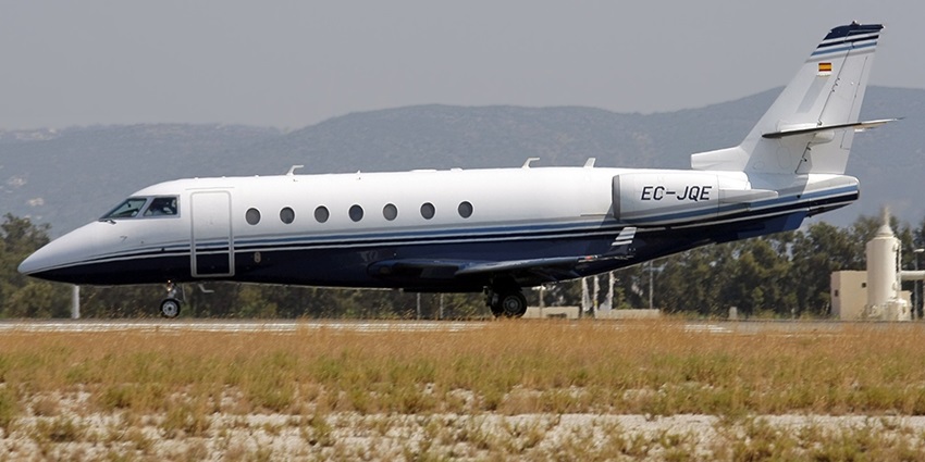 An aeroplane parked on a runway with mountains in the background and a dry, patchy grass field in front