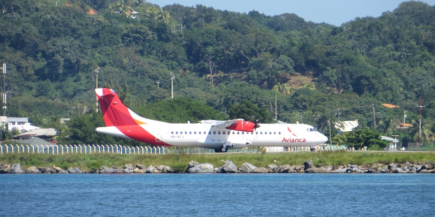 Avianca ATR-72 taxiing at Roatán Airport, surrounded by lush green hills and waters.