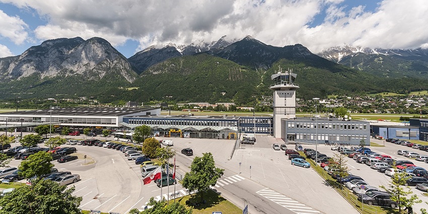 Daytime image of an airport surrounded by snow-clad mountains and cars in the parking space.