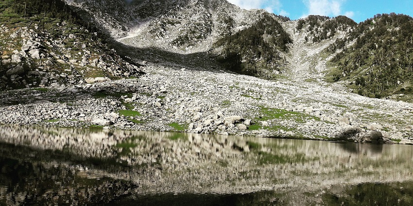 Kareri lake with a perfect reflection of rocky shoreline and mountains covered in clouds