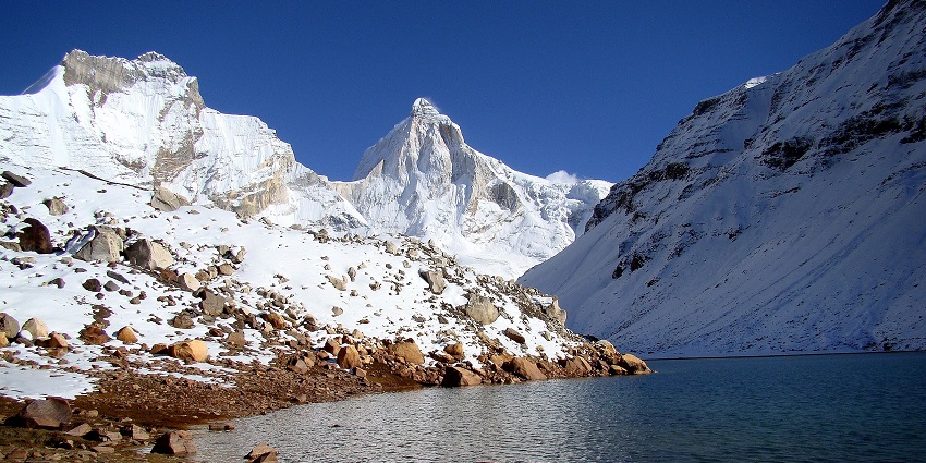 edar Tal with a clear water body, rocky shoreline and Thalay sagar peak covered in snow