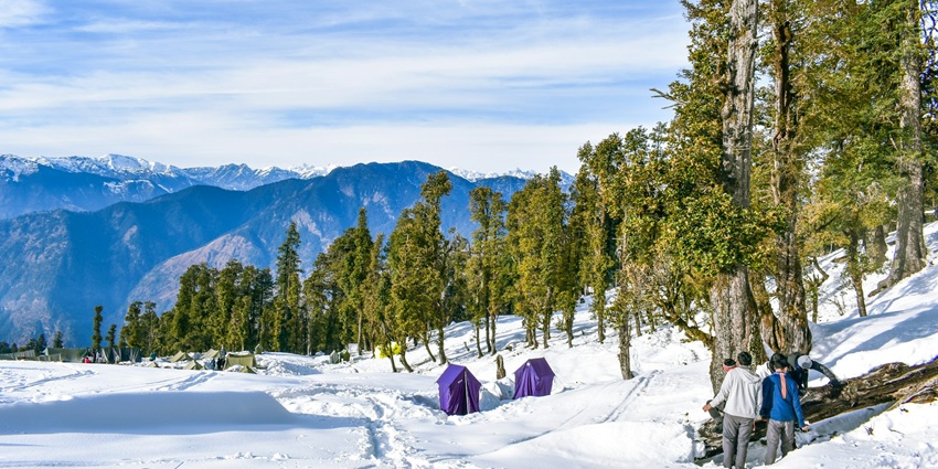 An image of a group standing in the snow in the Kedarkantha Trek, holding a sign marking Kedarkantha Summit.