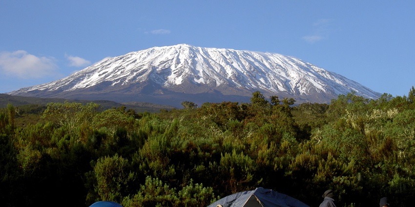 Mount Kilimanjaro’s snowy peak, green slopes, clear blue skies, tents, and climbers.