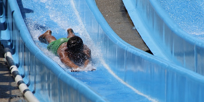 A photo of a boy sliding down the bright blue water slide into a clear turquoise swimming pool.