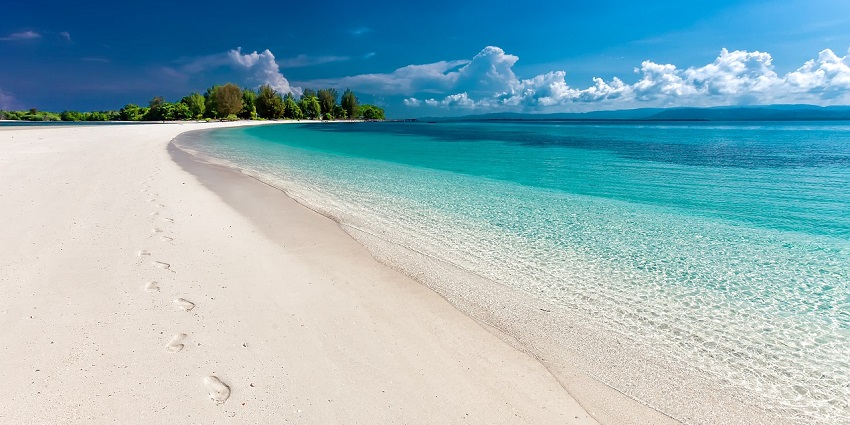 Golden sand meets the crystal clear blue waves under a sunny sky with white clouds.