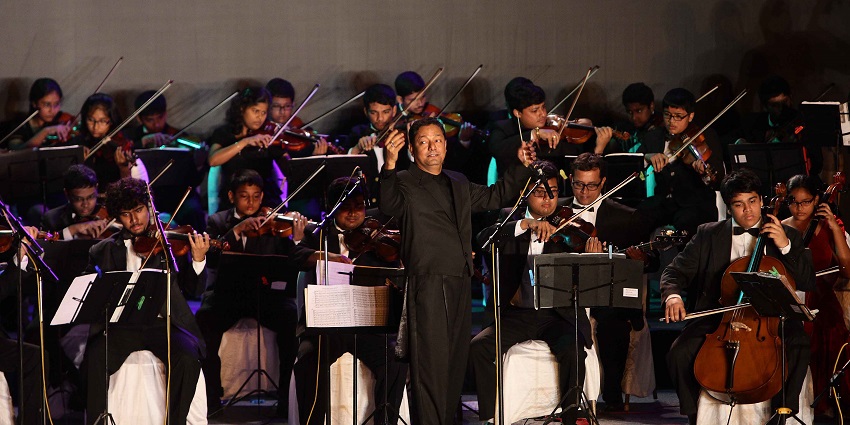 Orchestra performing on stage, with a conductor leading musicians playing string instruments.