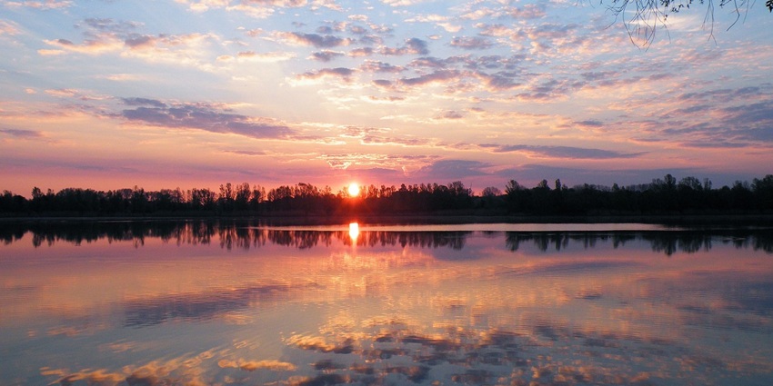 Stunning sunset over a lake with silhouetted trees and water reflecting the clouded sky.