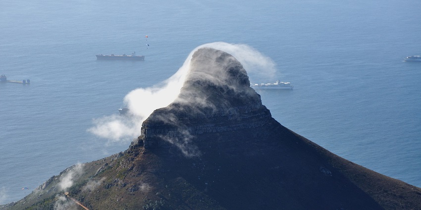 An image of the Lion’s Head mountain peak near the ocean, veiled by flowing clouds, with ships nearby.