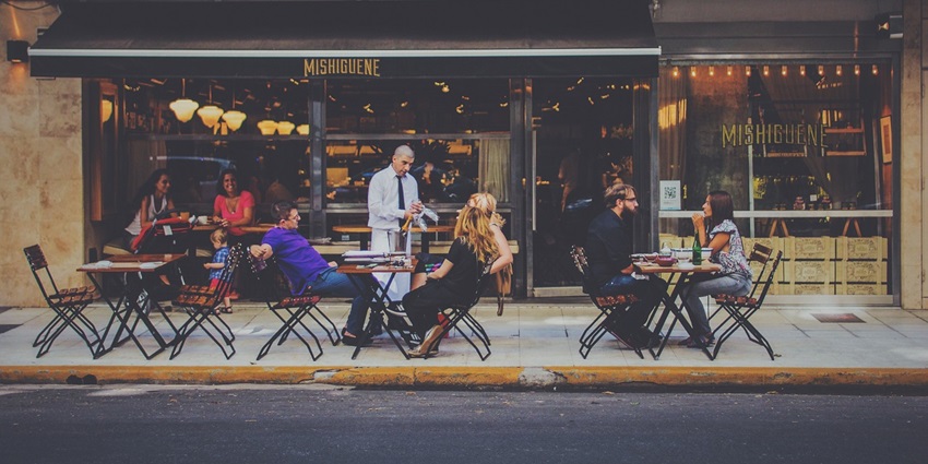 People are dining at an outdoor café while a waiter is serving drinks to customers.