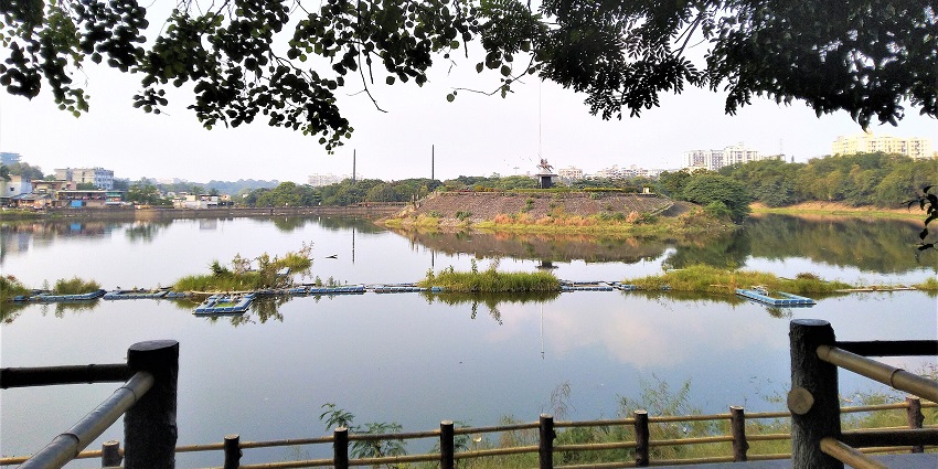 Upper Katraj Lake with calm water reflecting the sky and distant buildings offshore.