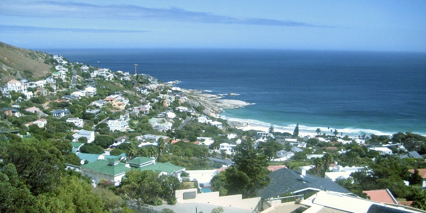 An image of the lively coastal area of Camps Bay Beach, with palm trees, a beach, bustling street, and mountain backdrop.