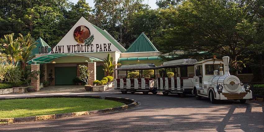 An image of Lok Kawi Wildlife Park's entrance with a triangular roof, many plants, and a small train