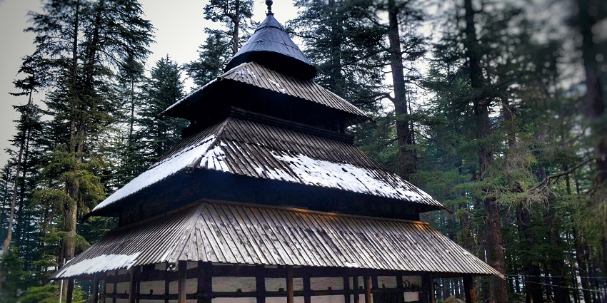 An image of the traditional wooden temple of Hadimba, with a multi-tiered pagoda-style roof, partially covered in snow in Manali.