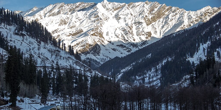 An image of a snowy mountainous landscape of Manali, with a multi-story building in the foreground, surrounded by trees.